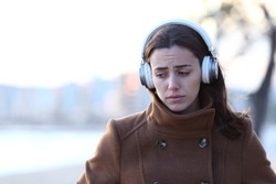 Front view of a sad woman walking alone wearing headphones listening to music in winter on the beach
