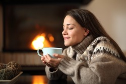 Happy woman in winter drinking coffee beside a fireplace heating at home