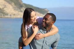 Funny interracial couple joking on piggyback on the beach on summer