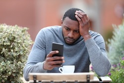 Front view portrait of a worried man with black skin checking phone in a coffee shop