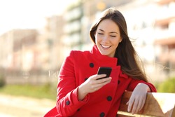 Girl texting on the smart phone sitting in a park wearing a red jacket and sitting in a bench in a park