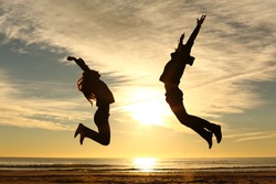 Couple or friends silhouette jumping on the beach at sunset with a warmth light and the sun in the middle