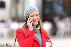 Happy woman calling on the mobile phone and holding a take away coffee in the street in winter