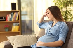 Pregnant woman with hand on forehead suffering headache sitting on a couch in the living room at home