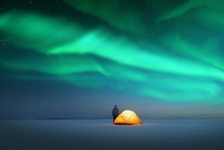 Tourist near yellow tent lighted from the inside against the backdrop of incredible starry sky with Aurora borealis. Amazing night landscape. Northern lights in winter field