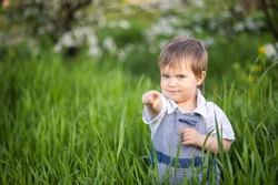 A little boy in denim overalls with expressive blue eyes. Jumping and fooling around in the tall green grass against the backdrop of a large green bush and a blooming home garden.