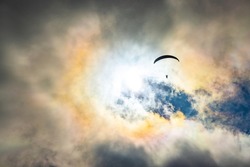 Paragliding adventure sport with parachute flying against bright sun hiding behind white cumulus clouds on blue sky on autumn day