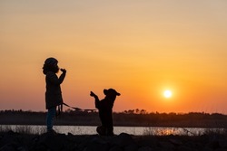An attentive, strict young girl in a warm beige jacket walks and trains her large obedient active guard dog of the Rottweiler breed against the backdrop of a small calm lake and a bright golden sunset