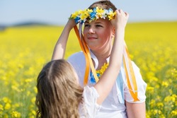 Kind cheerful younger sister puts on older smiling teenage brother bright Ukrainian flower wreath with long multi-colored ribbons, against background of blooming yellow boundless fields and clear sky