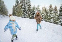 Side view of two children in outerwear throwing snowballs, playing in snowy forest at daytime. Happy female kids making snowballs, smiling, running in winter mountains. Concept of winter holidays.