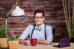 Strict boyish woman in suspenders at desk with a red coffee cup