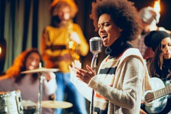 Close up of mixed race woman singing. In background band playing instruments. Home studio interior.