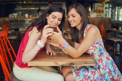 Two girls in a coffee shop, smiling, gossiping, drinking coffee, looking at smartphone 