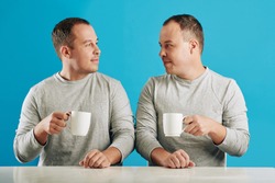 Young adult male twin siblings sitting at table looking at each other holding cups with coffee, blue background