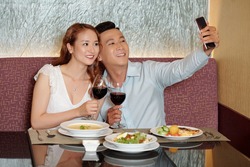 Smiling young couple taking selfie with wine glasses at table in fancy restaurant when having romantic dinner