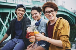 Group portrait of smiling Vietnamese friends looking at camera while toasting with soft drink bottles