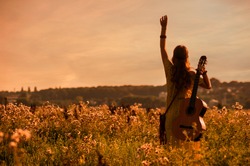 silhouette of woman wearing a bohemian style holding a guitar on a field at warm light of sunset