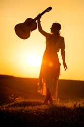silhouette of woman wearing a bohemian style holding a guitar on a field at warm light of sunset