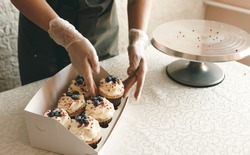 A girl cook in a gray apron packs cupcakes with cream in a gift box to send the order to the customer.