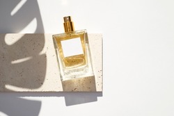 Transparent bottle of perfume with white label on stone plate on a white background. Fragrance presentation with daylight. Trending concept in natural materials with palm leaf shadows.