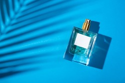 Transparent bottle of perfume with label on a blue background. Fragrance presentation with daylight. Trending concept in natural materials with palm leaves shadow. Women's and men's essence.