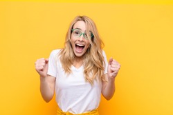 young pretty blonde woman feeling shocked, excited and happy, laughing and celebrating success, saying wow! against flat color wall
