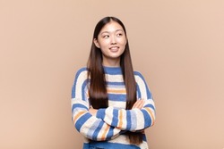 asian young woman feeling happy, proud and hopeful, wondering or thinking, looking up to copy space with crossed arms