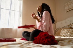 happy african american woman eating romantic valentines chocolates from heart shaped box while sitting on bed at home