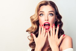Shocked and surprised girl screaming covering  mouth her hands.Curly hair woman amazed.Beautiful girl  with curly hairstyle and red nails manicure.Presenting your product.Expressive facial expressions
