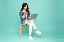 Young happy smiling Asian woman holding laptop computer and sitting on chair isolated on green background, Chatting and laughing with friends online concept