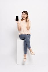 Happy Asian woman sitting and showing mobile phone and mini heart sign isolated over white background