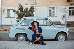 Pretty relaxing woman in hat sitting near vintage blue car. Lady with cup of drink. Leisure time, coffee break concept