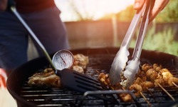 A food thermometer checking that chicken is being cooked correctly, safely on a barbecue on a sunny day in the garden as a man turns over kebabs with tongs.