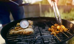 A food thermometer checking that chicken is being cooked correctly, safely on a barbecue as a man turns over prawns with a pair of tongs on a sunny day in the garden.
