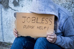 Jobless man begging on a street holding a handwritten sign for help covering his face as he sits on the sidewalk in jeans and a hoodie holding a cup for coins