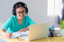 Asian young woman student with glasses headphones girl study happy writing note on a book looking video conference laptop computer university class online internet learning distance education at home