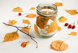 Autumn-themed DIY candle. Tealight candle in glass jar, decorated dried golden leaves. Fall time interior decor. 