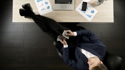Relaxed wealthy businessman counting money, sitting with feet on table, top view