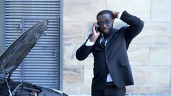 Businessman standing by broken car with open hood, talking over phone, breakdown