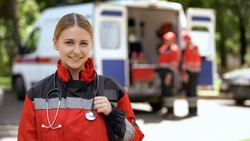 Female paramedic smiling into camera, ambulance crew blurred on background
