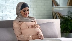 Smiling pregnant lady in hijab holding tummy, baby expectation, motherhood