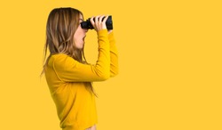 young girl with yellow sweater and looking in the distance with binoculars on isolated yellow background