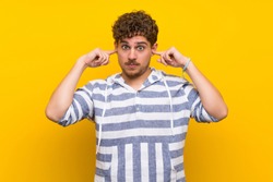 Blonde man over yellow wall covering both ears with hands