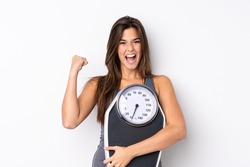 Teenager Brazilian girl holding a scale over isolated white background with weighing machine and doing victory gesture
