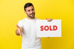 Young handsome caucasian man isolated on yellow background holding a placard with text SOLD making a deal