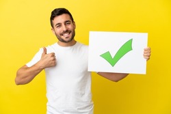 Young handsome caucasian man isolated on yellow background holding a placard with text Green check mark icon with thumb up