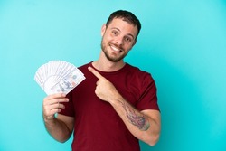 Young Brazilian man taking a lot of money over isolated background pointing to the side to present a product