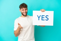 Young handsome caucasian man isolated on blue background holding a placard with text YES making a deal