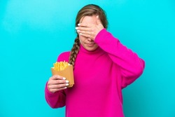 Young caucasian woman holding fried chips isolated on blue background covering eyes by hands. Do not want to see something