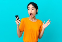 Young Vietnamese woman isolated on blue background looking at the camera while using the mobile with surprised expression